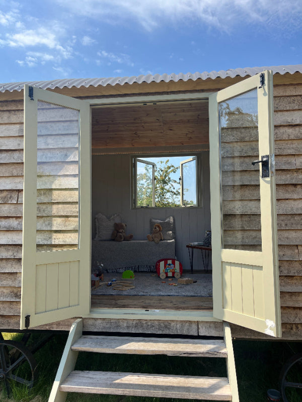A large wooden shepherd’s hut with open French doors leading up a set of steps.